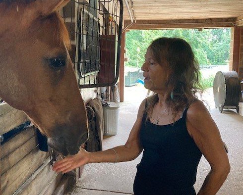 Donna Sue Rosenthal in a barn aisle, hand extended to a chestnut horse leaning out of its stall